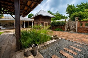Une maison traditionnelle en bois, avec un toit en tuiles, entourée d’un jardin où poussent différentes espèces de plantes et bordée d’un mur en pierre.