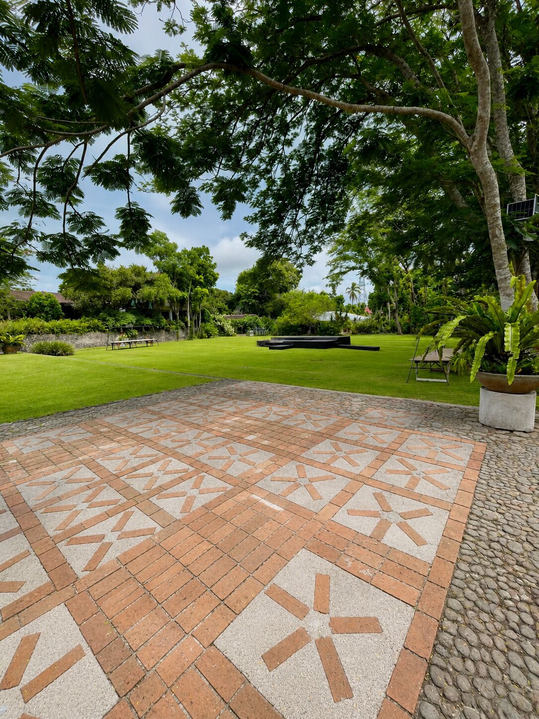 Eine gepflasterte Terrasse mit geometrischem Muster, umgeben von üppigem Grün und Bäumen.