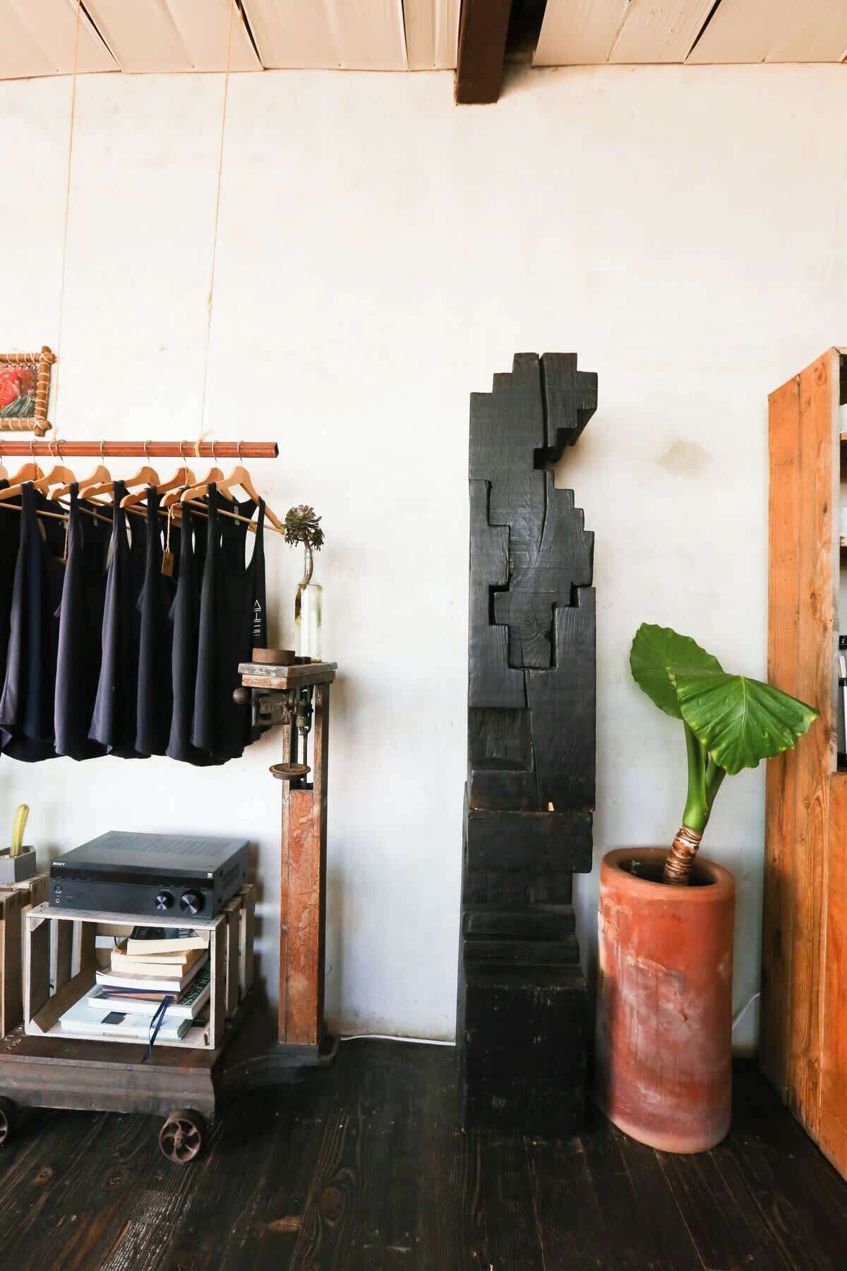 A room with a wooden floor, a black abstract sculpture, a potted plant, a clothing rack with dark garments, and a vintage Sony stereo system.