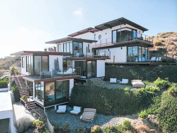 Une maison moderne à plusieurs étages, dotée de grandes fenêtres en verre, d’un espace extérieur aménagé et offrant une vue sur le paysage environnant.