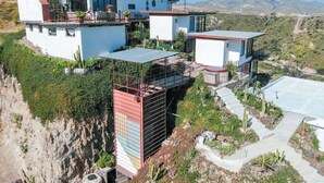 Uma casa moderna de vários andares com telhado plano, grandes janelas e varanda com vista para uma paisagem desértica com montanhas ao fundo.