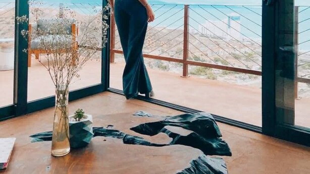 Uma mulher em pé perto de uma grande janela com vista para a paisagem costeira.