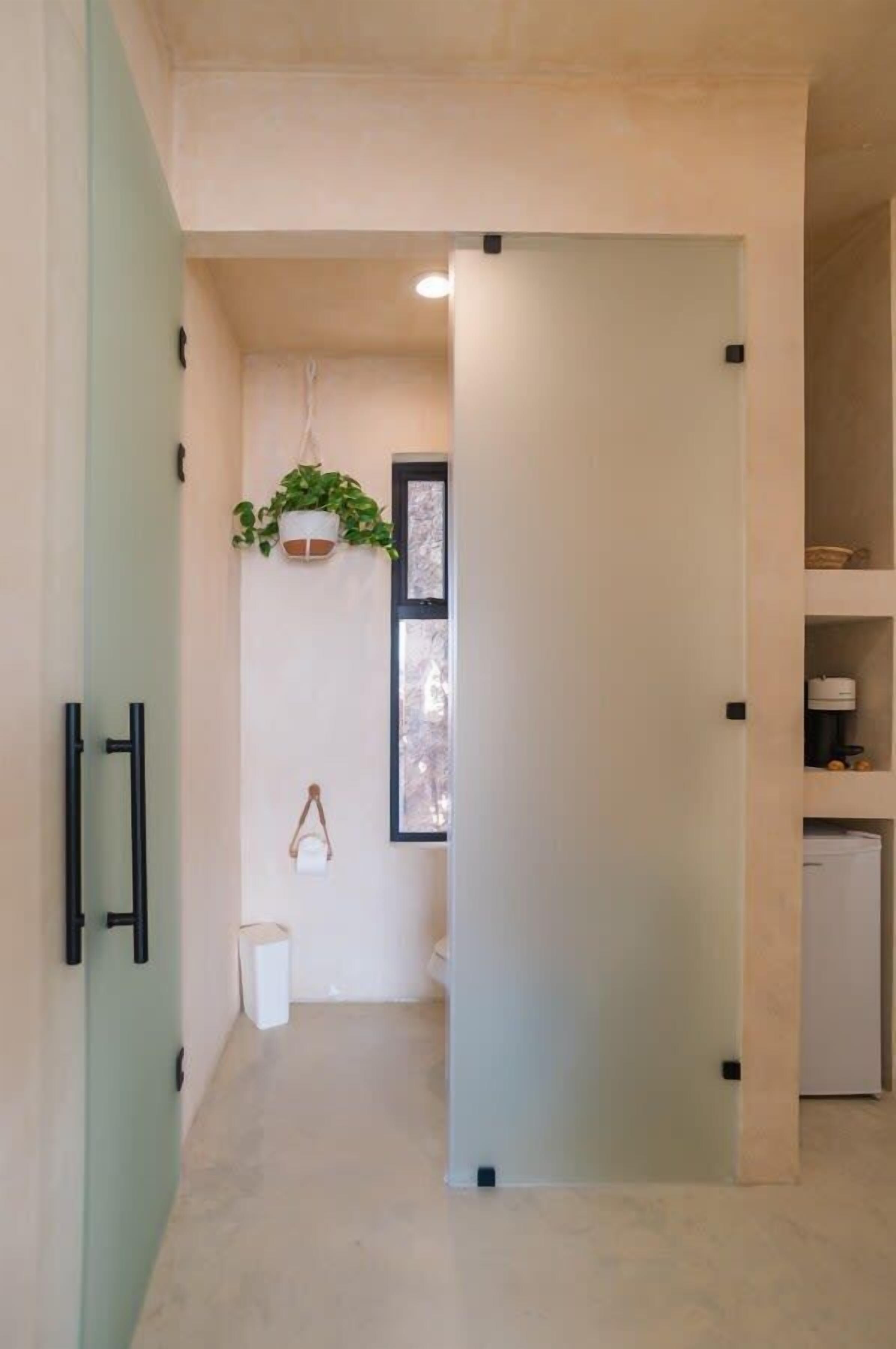 A narrow hallway with a door leading to a room. A potted plant hangs on the wall, and a small white trash bin is visible.