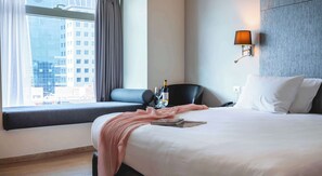 Une chambre d’hôtel avec un grand lit, une fenêtre donnant sur des immeubles, un bureau avec une chaise et une lampe fixée au mur.