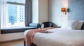Ein Hotelzimmer mit einem großen Bett, einem Fenster mit Blick auf Gebäude, einem Schreibtisch mit Stuhl und einer Wandlampe.