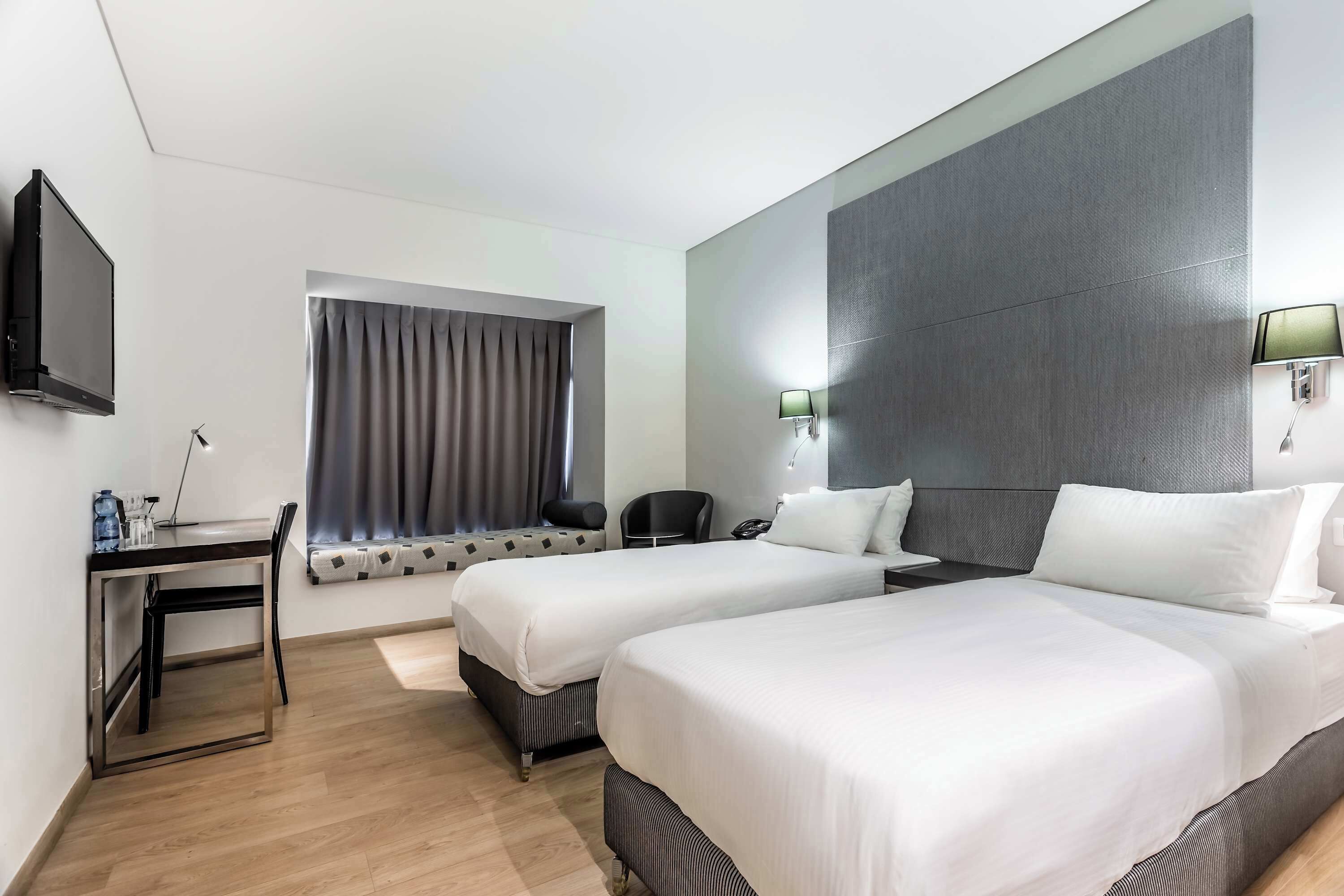 A hotel room with two beds, a desk, a chair, a TV, and a window with curtains.