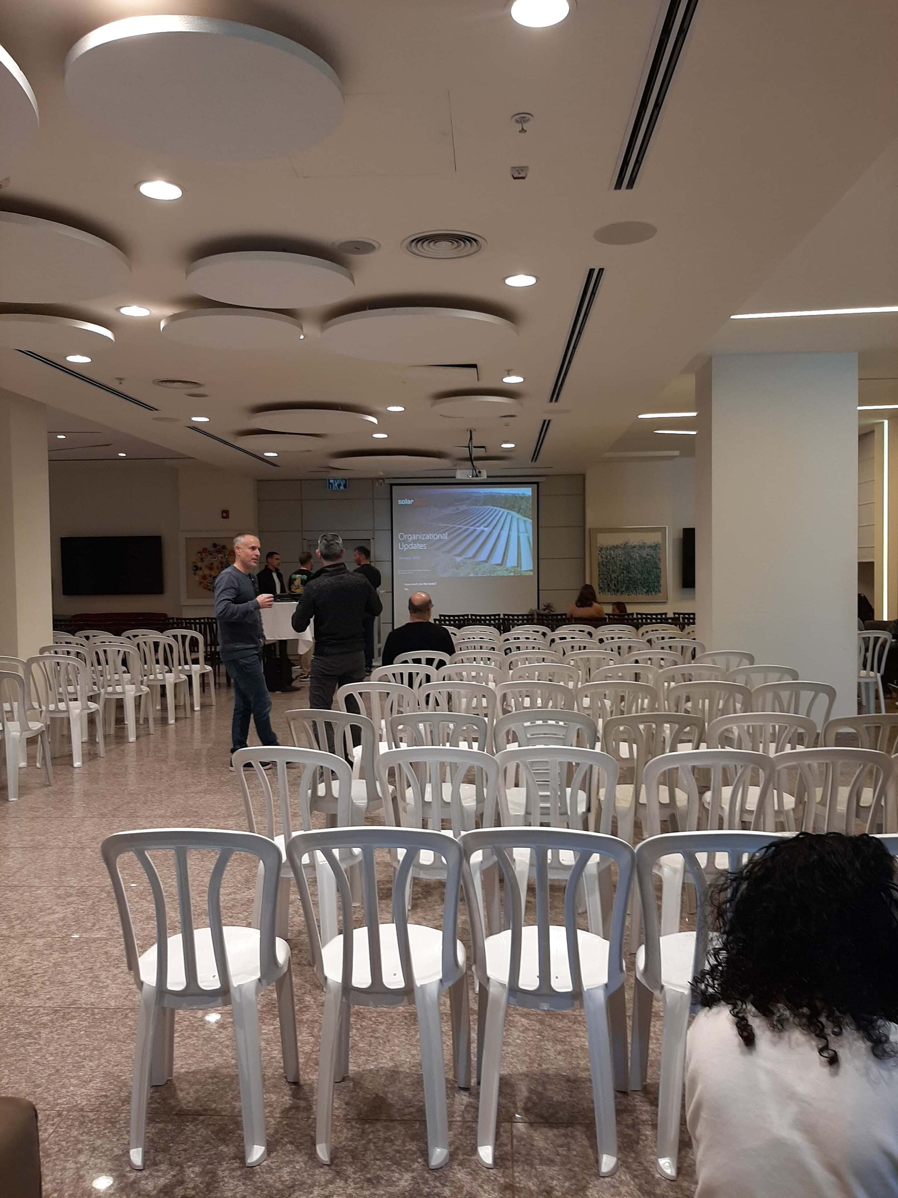 A conference room with rows of white plastic chairs, a large screen displaying a presentation, and a modern ceiling with recessed lighting.