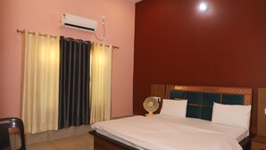 A bedroom with a bed, a bedside table, a chair, an air conditioner, and a window with curtains. - Royal Chuka Farm Stay (Puranpur)