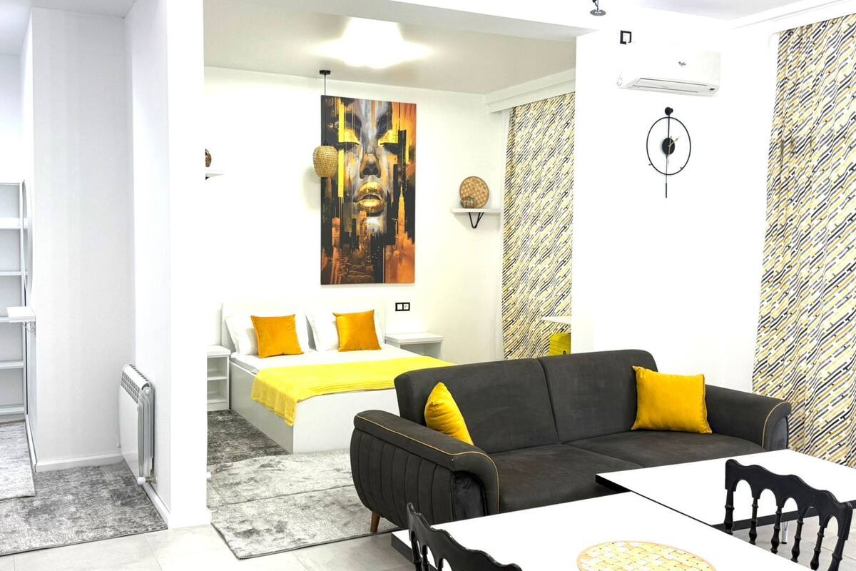 Een moderne woonkamer met een bank, een eettafel en een bed met gele kussens.