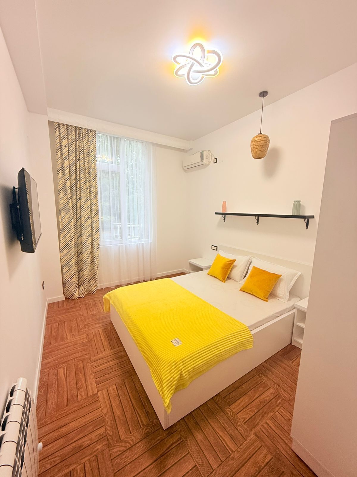 A bedroom with a bed, a television, an air conditioner, a window with curtains, a hanging light, and a shelf with items.