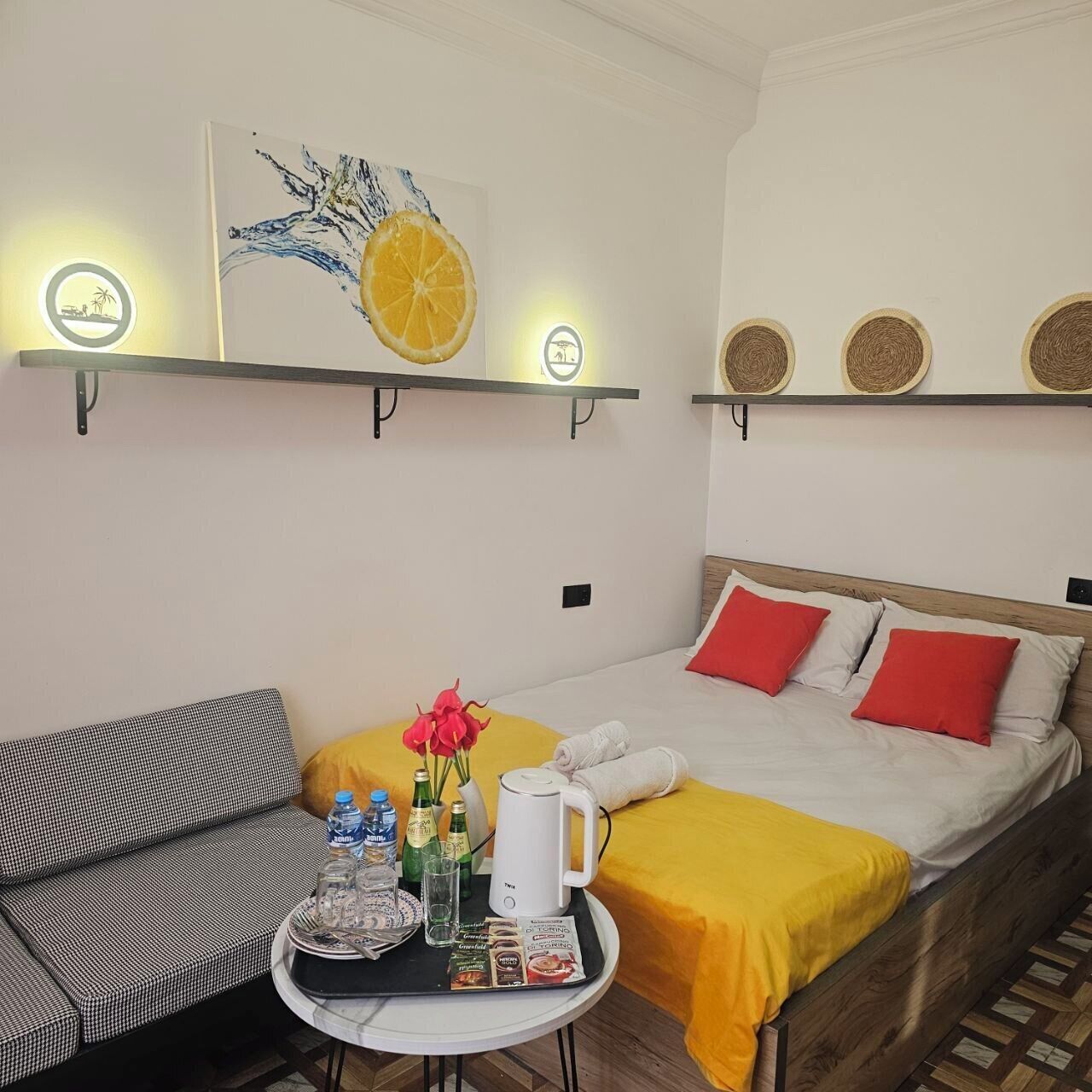Une pièce confortable avec un lit, un canapé, une petite table sur laquelle se trouve un plateau de produits pour le petit-déjeuner, et des étagères murales ornées d’objets décoratifs.