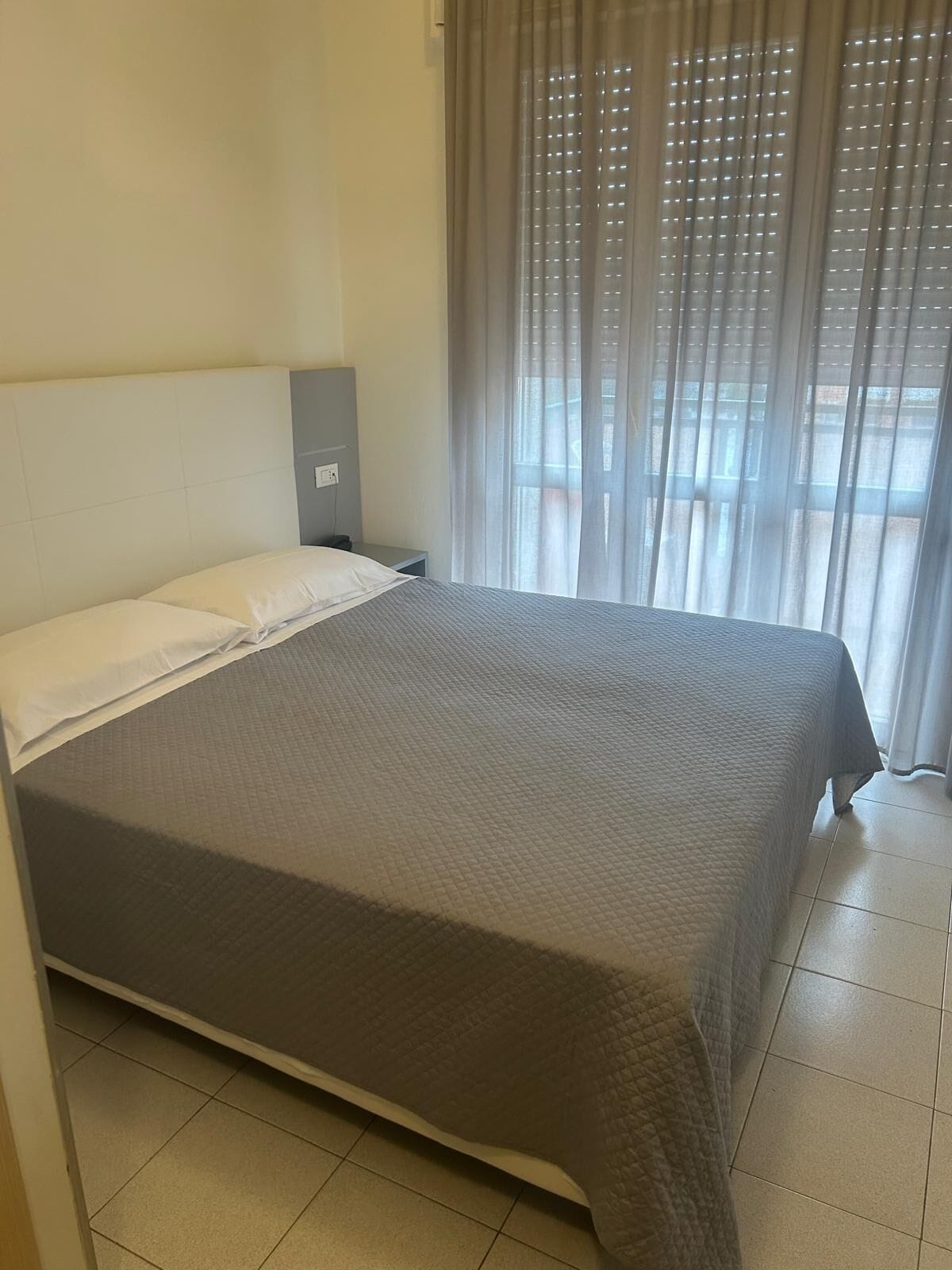 A neatly made bed with a grey quilt, a bedside table with a lamp, and a window with sheer curtains.