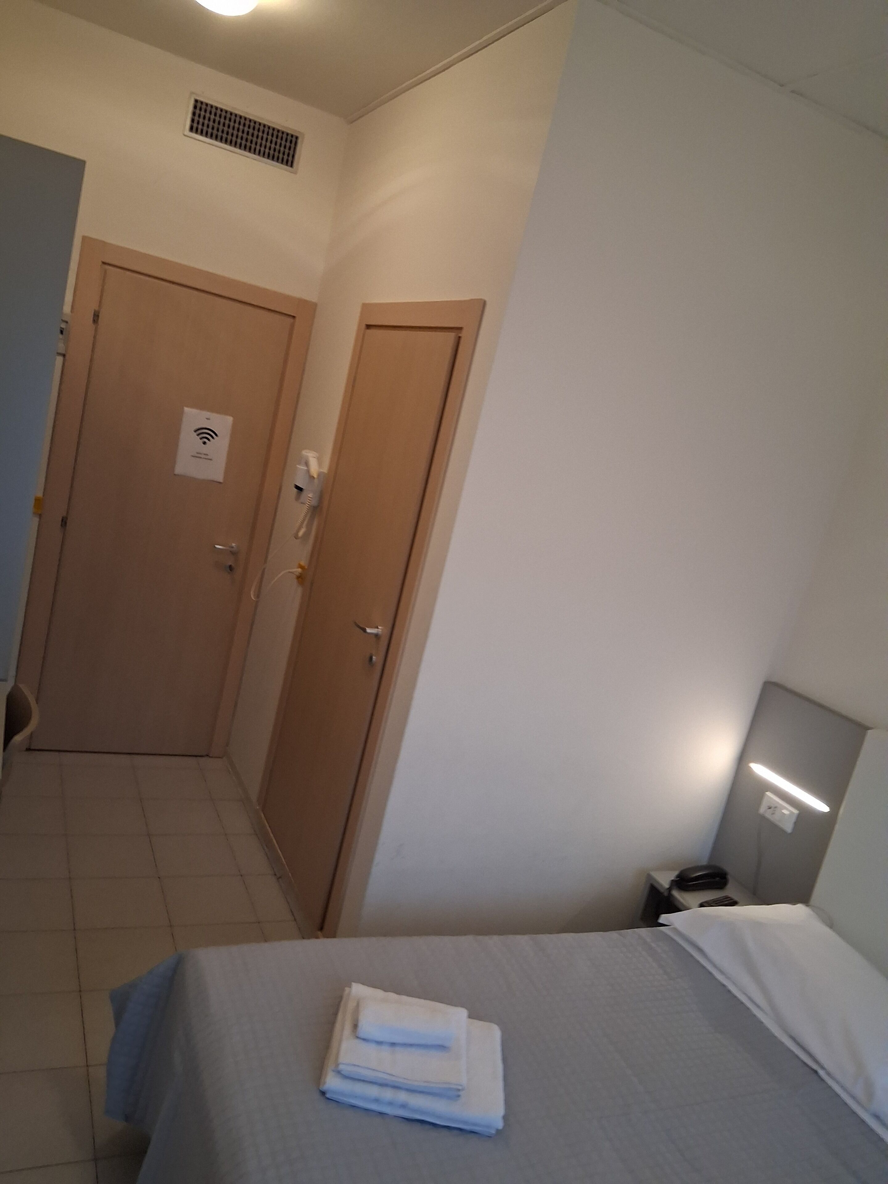 Ein Hotelzimmer mit einem Bett, einem Nachttisch, einer Lampe, einer Tür mit WLAN-Symbol und einem Lüftungsgitter an der Decke.