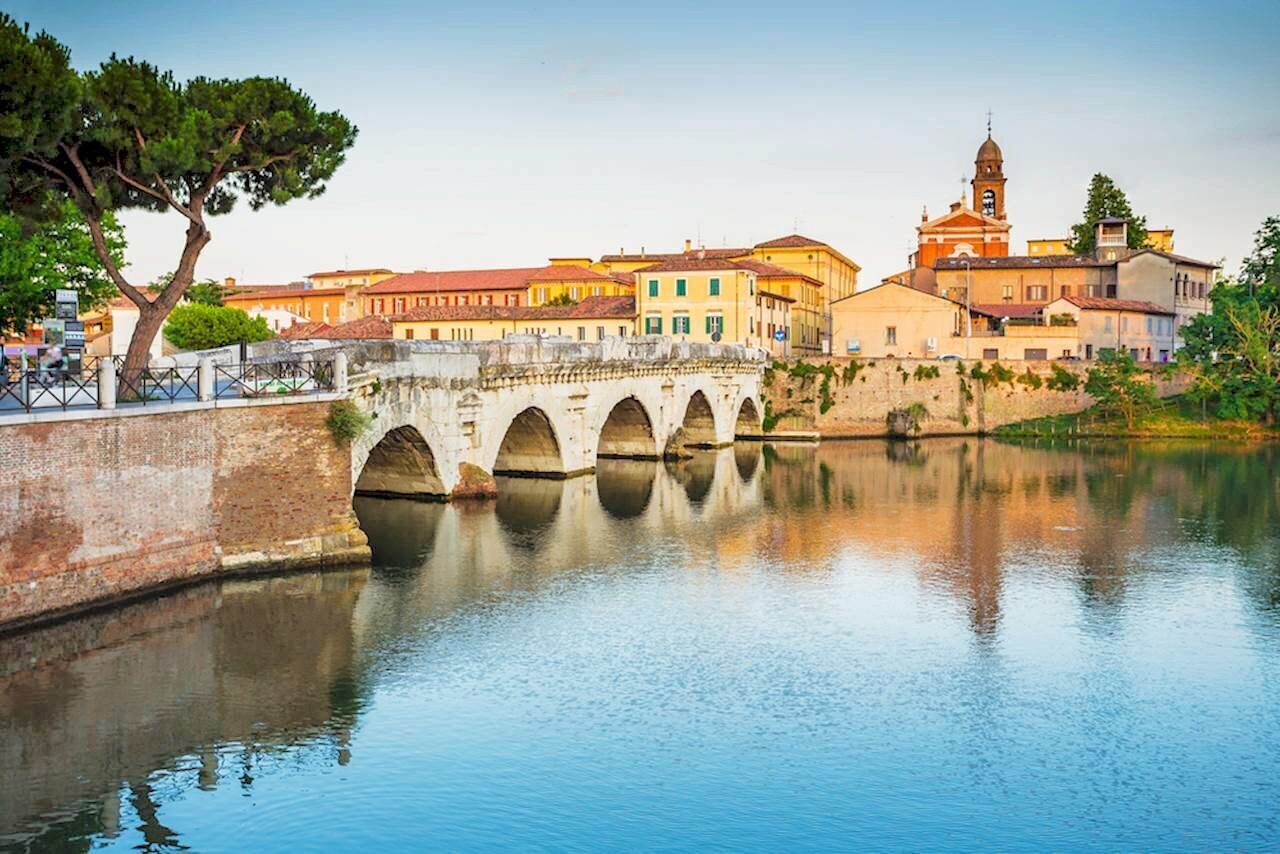 Un puente histórico sobre un río tranquilo, con edificios y una iglesia al fondo.