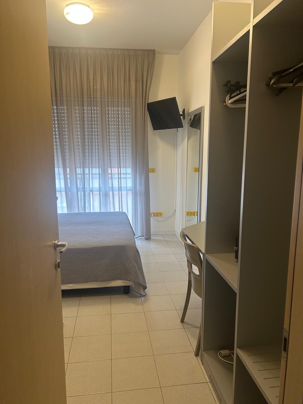A hotel room with a bed, a desk, a wardrobe, a TV mounted on the wall, and a window with curtains.