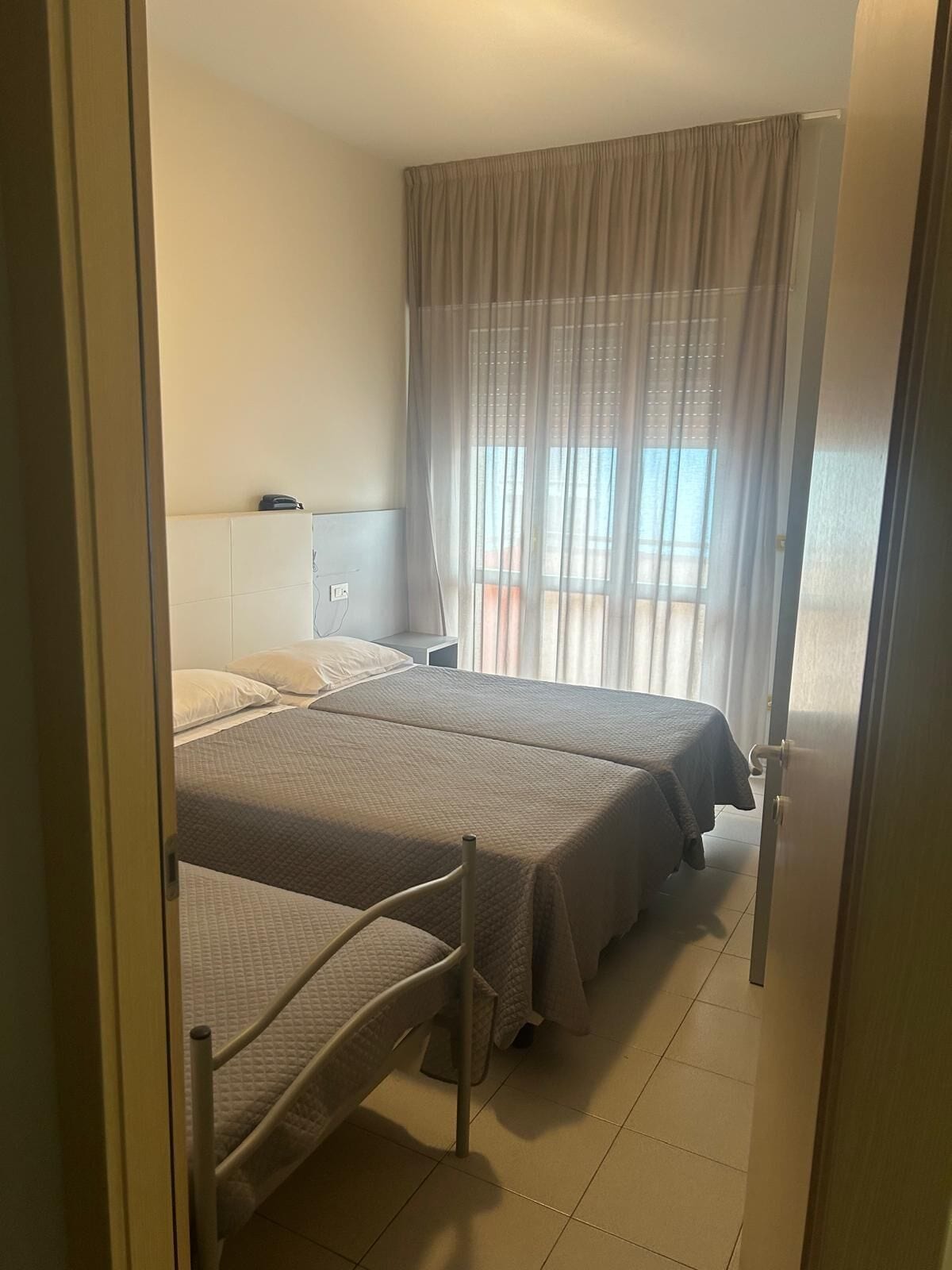 A hotel room with two beds, a bedside table, a window with sheer curtains, and a view of the outside.