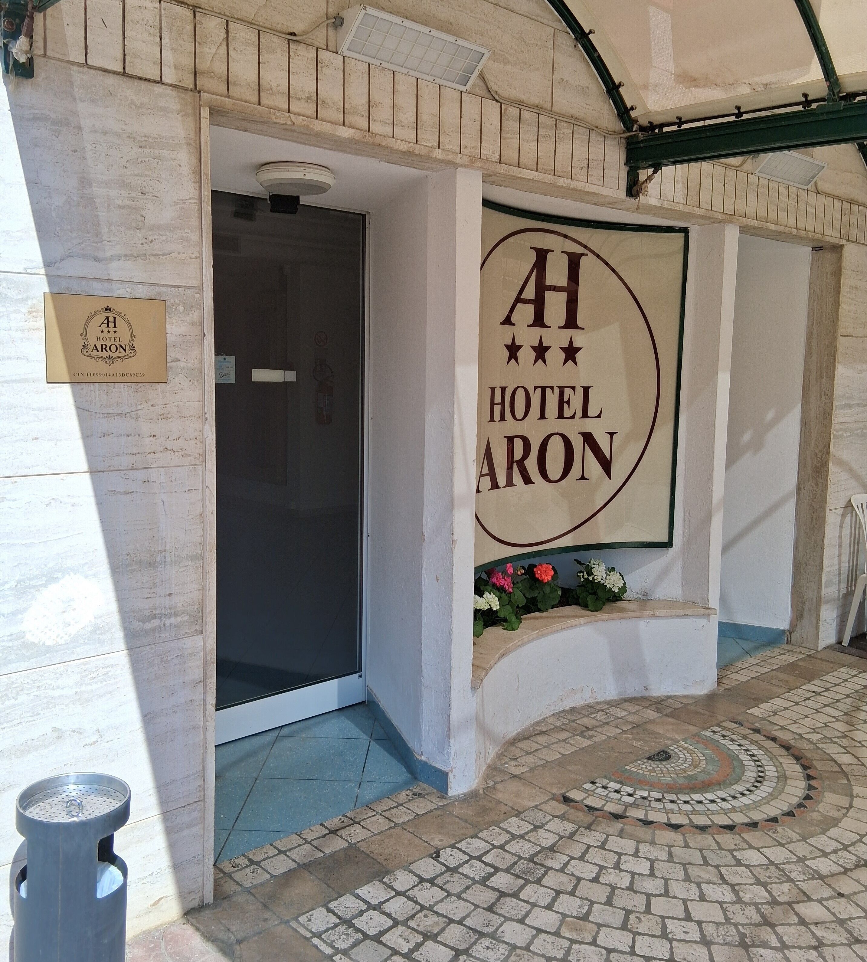 A hotel entrance with a sign reading 'AH HOTEL ARON,' a tiled entrance, and a small flower arrangement.