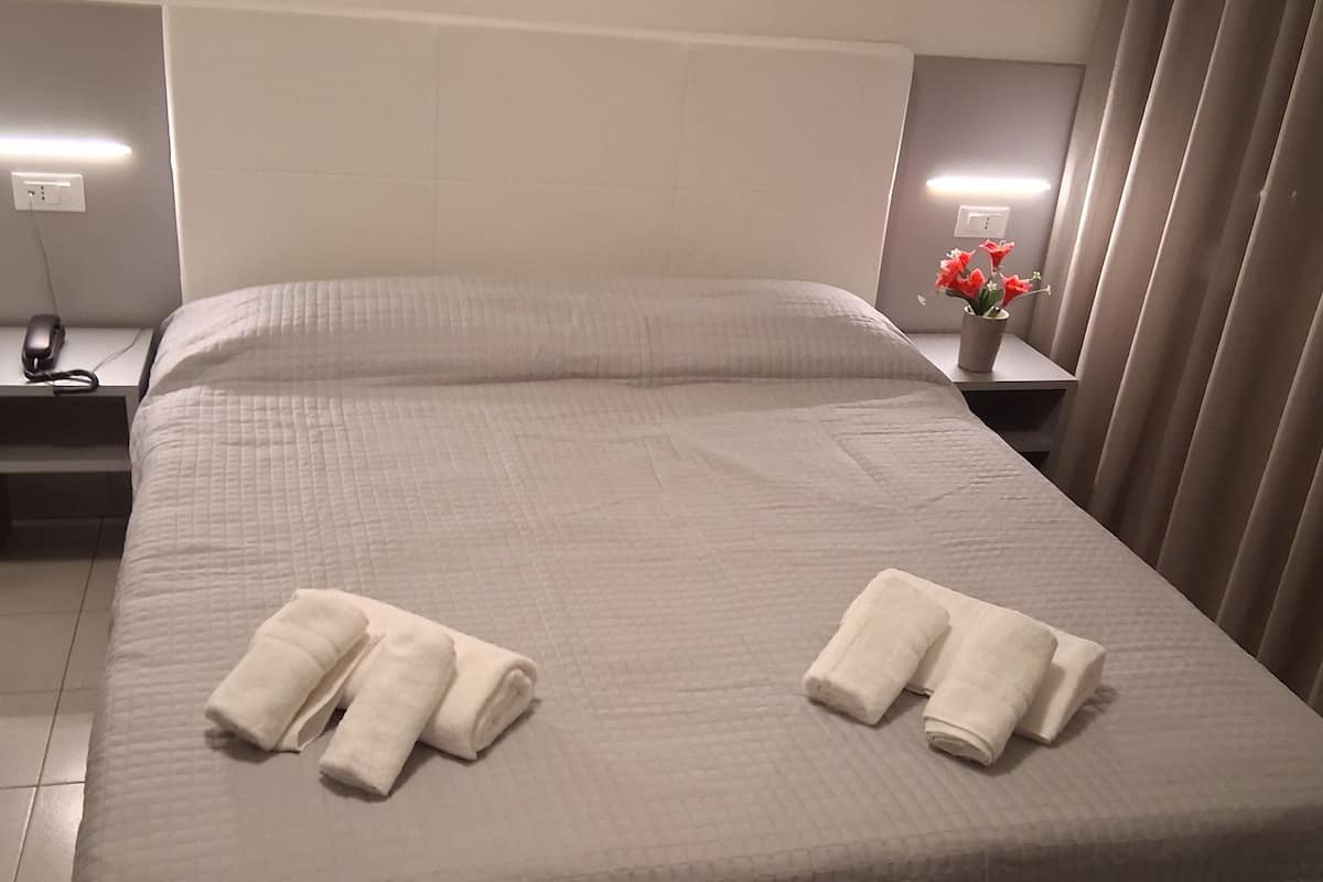 A neatly made bed with white linens and towels, a bedside table with a lamp, and a potted plant.