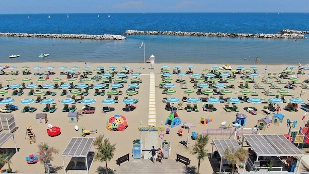 A beach with numerous sunbeds and umbrellas, a lifeguard tower, and a rocky breakwater in the background. - Hotel Aron Rimini (Rimini)