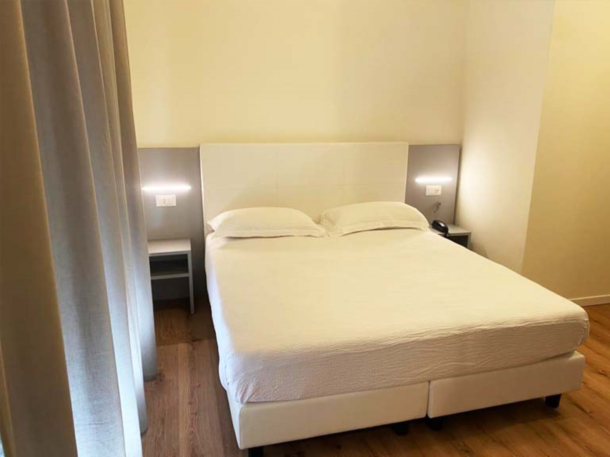 A single bed with a white headboard and footboard, a bedside table with a lamp, and a wooden floor.