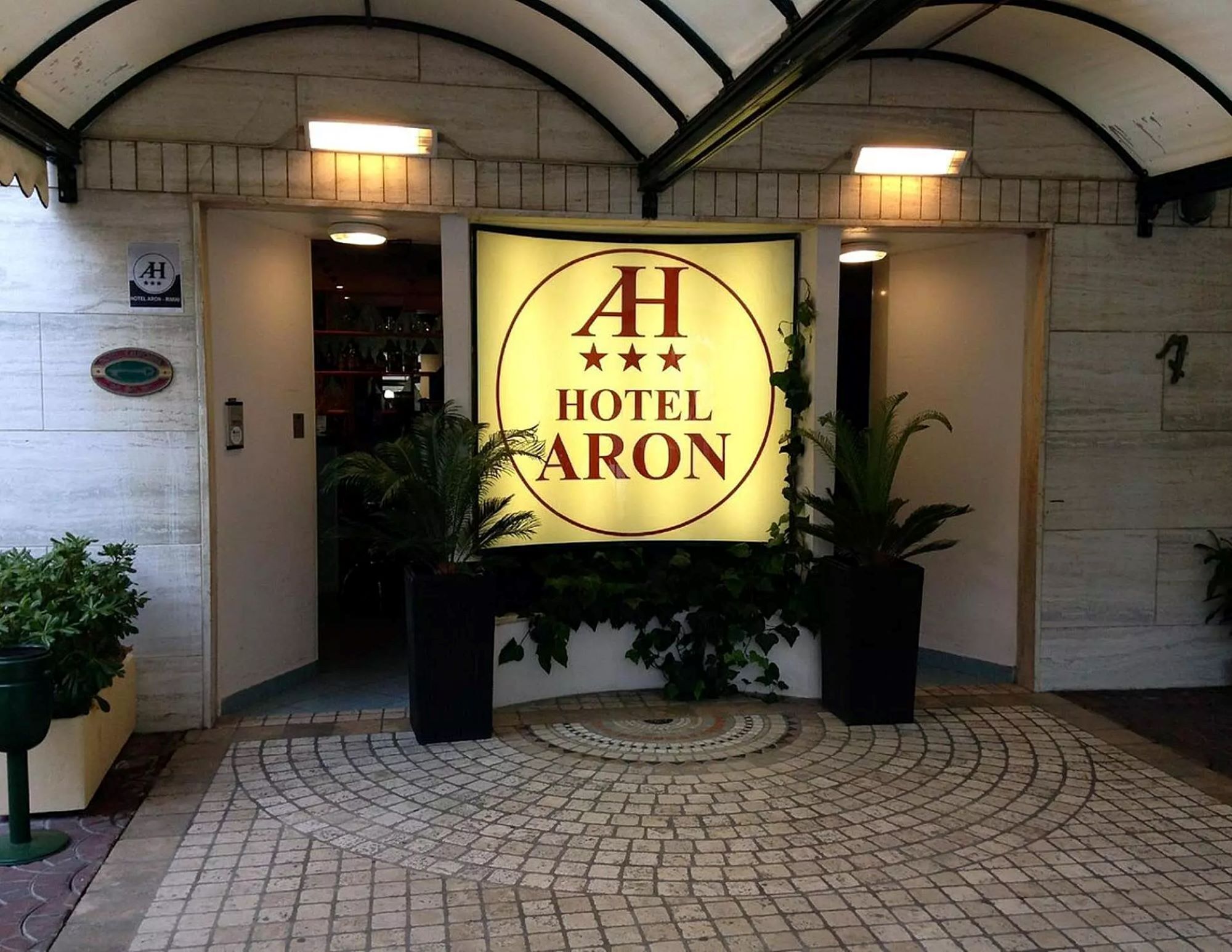 The entrance of Hotel Aron with a prominent illuminated sign, tiled flooring, and potted plants.