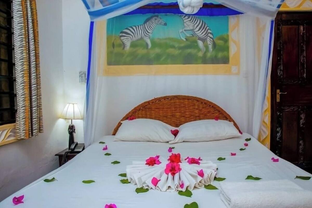 A neatly made bed with white linens, pink and green flower petals, and a folded towel centerpiece.