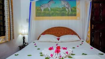 Una cama bien hecha con sábanas blancas, pétalos de flores rosa y verde, y un centro de mesa con una toalla doblada.