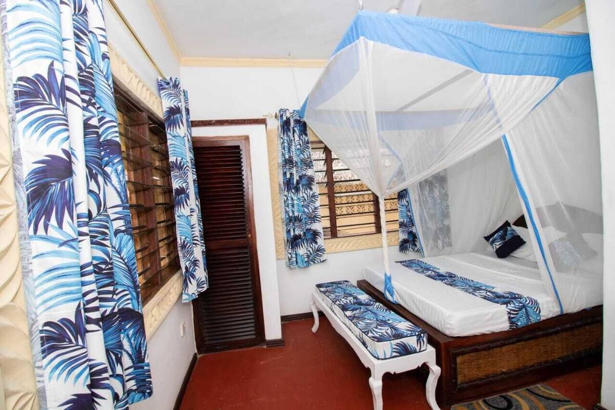 A bedroom with a bed under a mosquito net, a bench, and curtains with a leaf pattern.