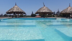 A swimming pool with clear water, surrounded by thatched umbrellas, and a red-roofed building in the background. - Malindi Breeze Point Hotel (Malindi)