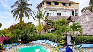 Un hôtel doté d’une piscine, de palmiers et d’un bâtiment à toit de chaume sur lequel est inscrit « TABLE DE BILLARD – SALLE DE CONFÉRENCE ».