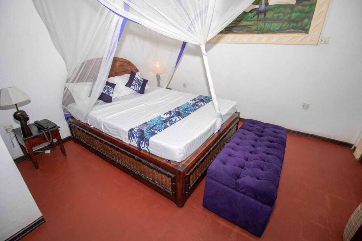 A bed with a canopy, a bedside table with a lamp, and a purple ottoman.