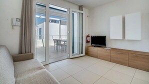 Une chambre d’hôtel avec une porte-fenêtre coulissante donnant sur un balcon, un canapé, un téléviseur posé sur un meuble en bois et un extincteur.