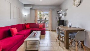 Un salon moderne avec un canapé d’angle rouge, une table à manger en bois avec des chaises, une petite table basse, un téléviseur et une horloge murale.