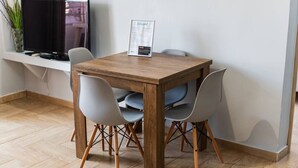 Un coin repas avec une table en bois et quatre chaises, un panneau posé sur la table, et un téléviseur placé sur une étagère au-dessus.