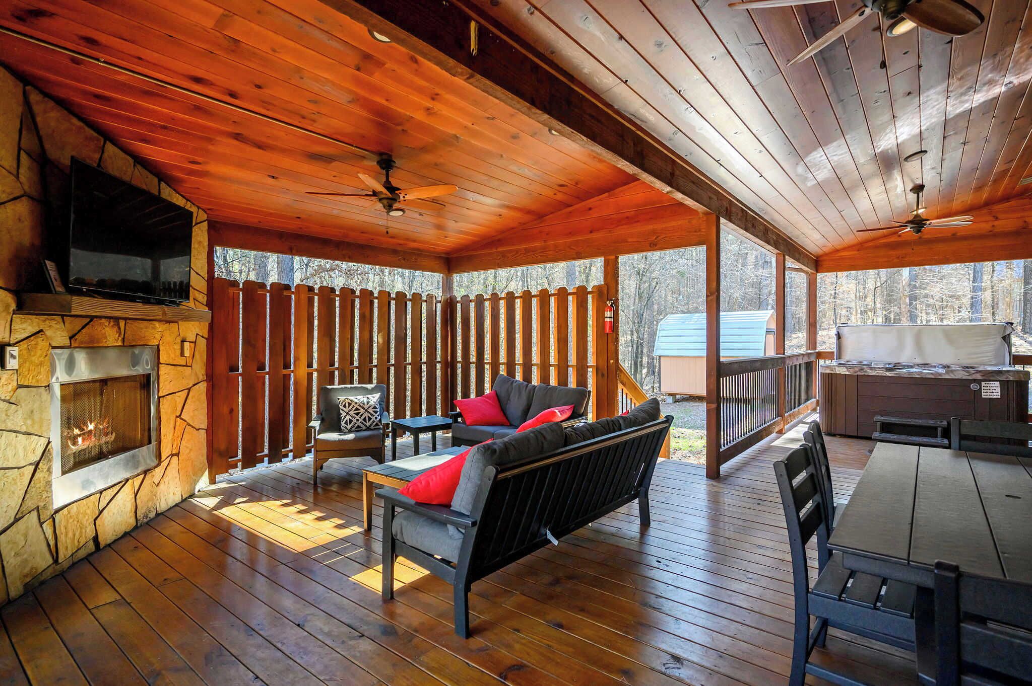 A covered outdoor area with a stone fireplace, wooden deck, seating, and a television.