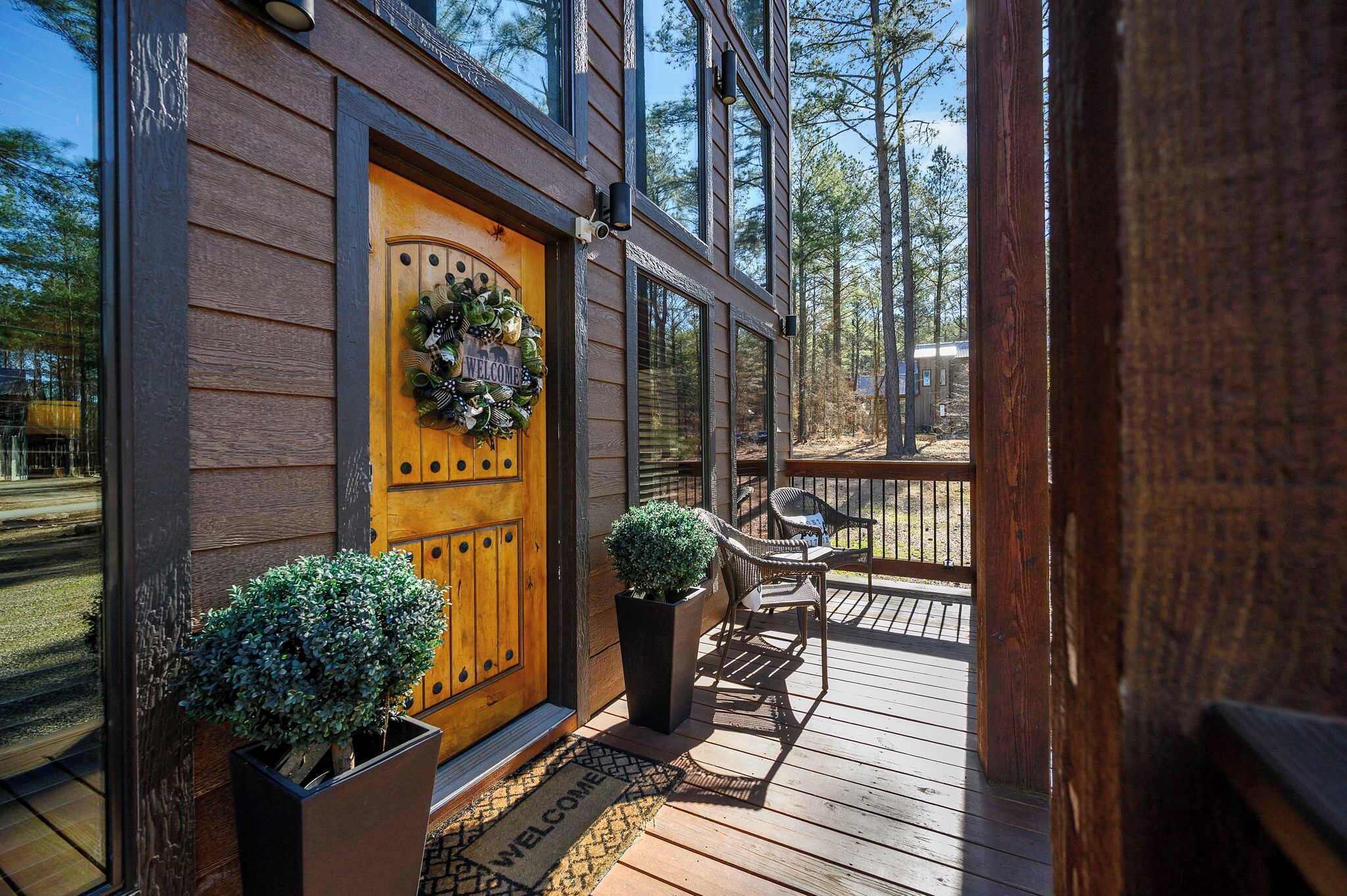 A wooden door with a welcome wreath, two potted plants, and a bench on a wooden deck.
