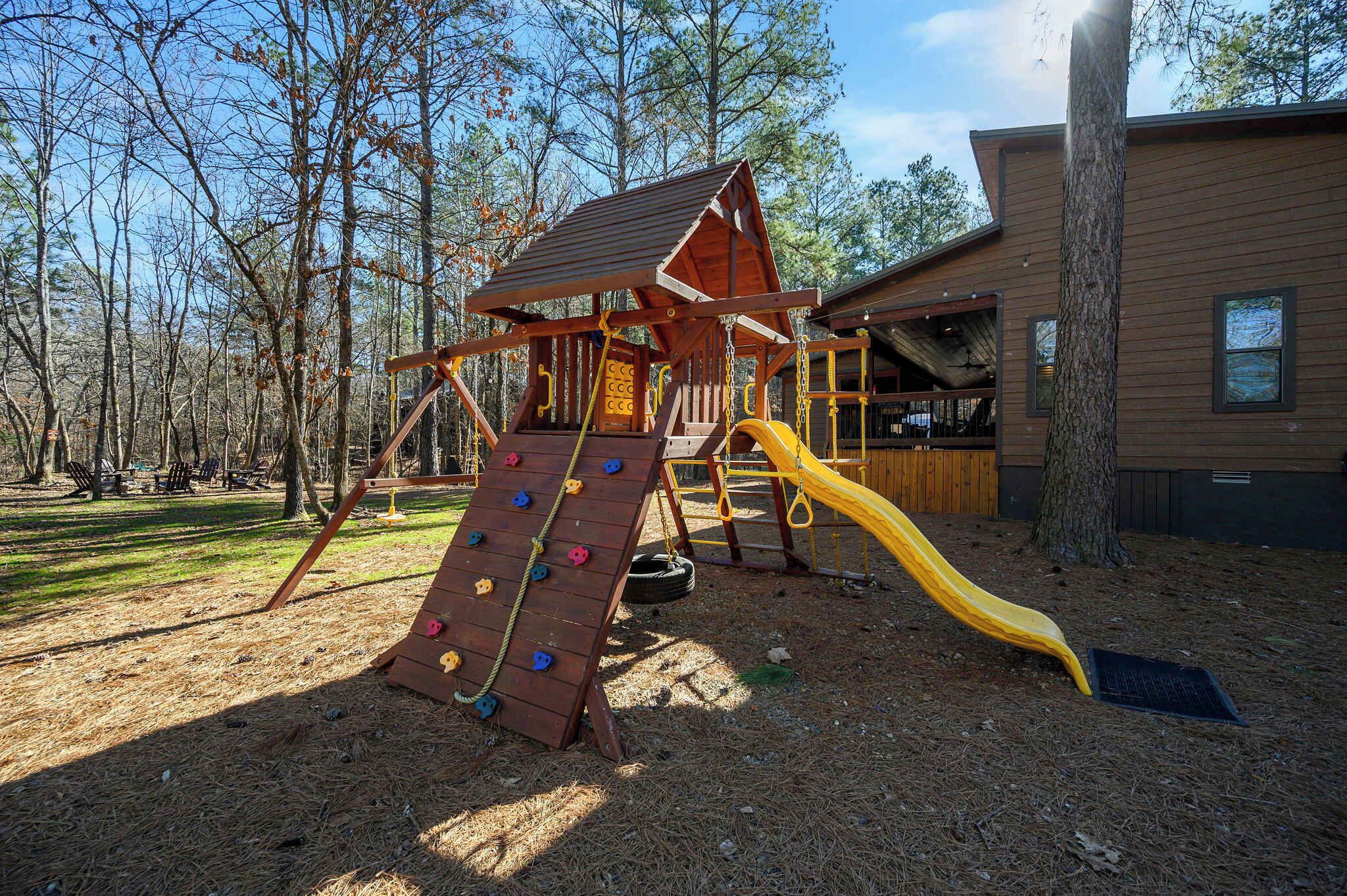 A playground with a climbing wall and slide, surrounded by trees and a wooden building.