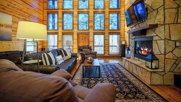 A cozy log cabin interior with a stone fireplace, a large TV mounted on the wall, and a view of the forest through the windows. - Hoot's Hideaway w/ Hot Tub (Broken Bow)
