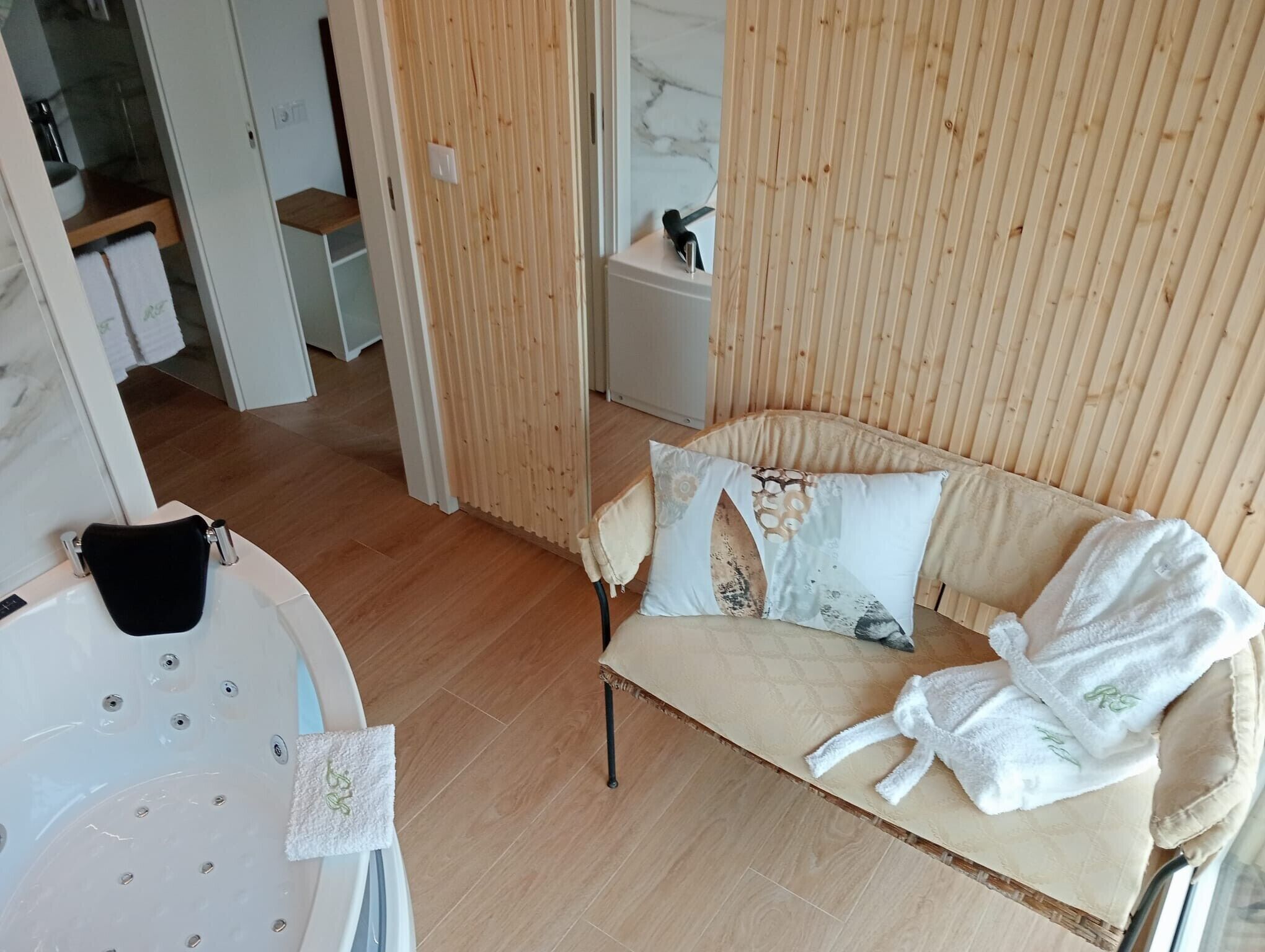 A room with a wooden partition, a sofa with patterned cushions, a white bath tub, and a robe on the sofa.