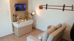 En moderne stue med en træpanelfront, et vægmonteret TV, en hvid skranke med dekorative genstande, en beige sofa og en træreol.
