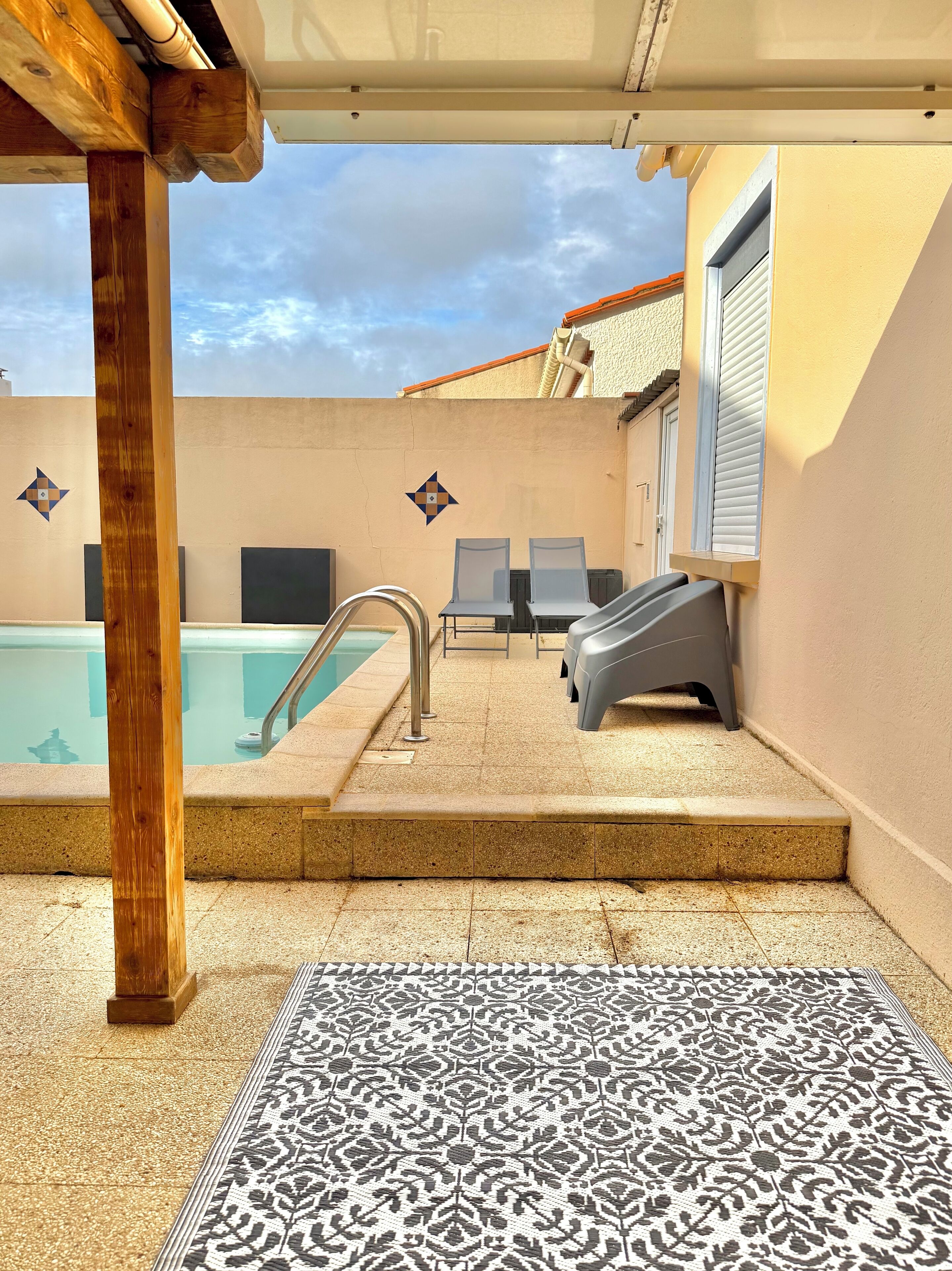 A pool area with a tiled floor, a pool, two lounge chairs, a small table, and a patterned rug.
