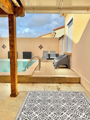 A pool area with a tiled floor, a pool, two lounge chairs, a small table, and a patterned rug.