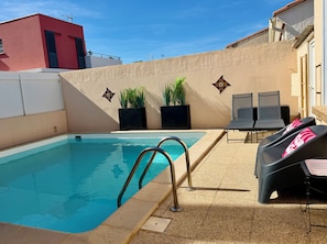 A swimming pool with a metal ladder, two lounge chairs, and potted plants against a wall.