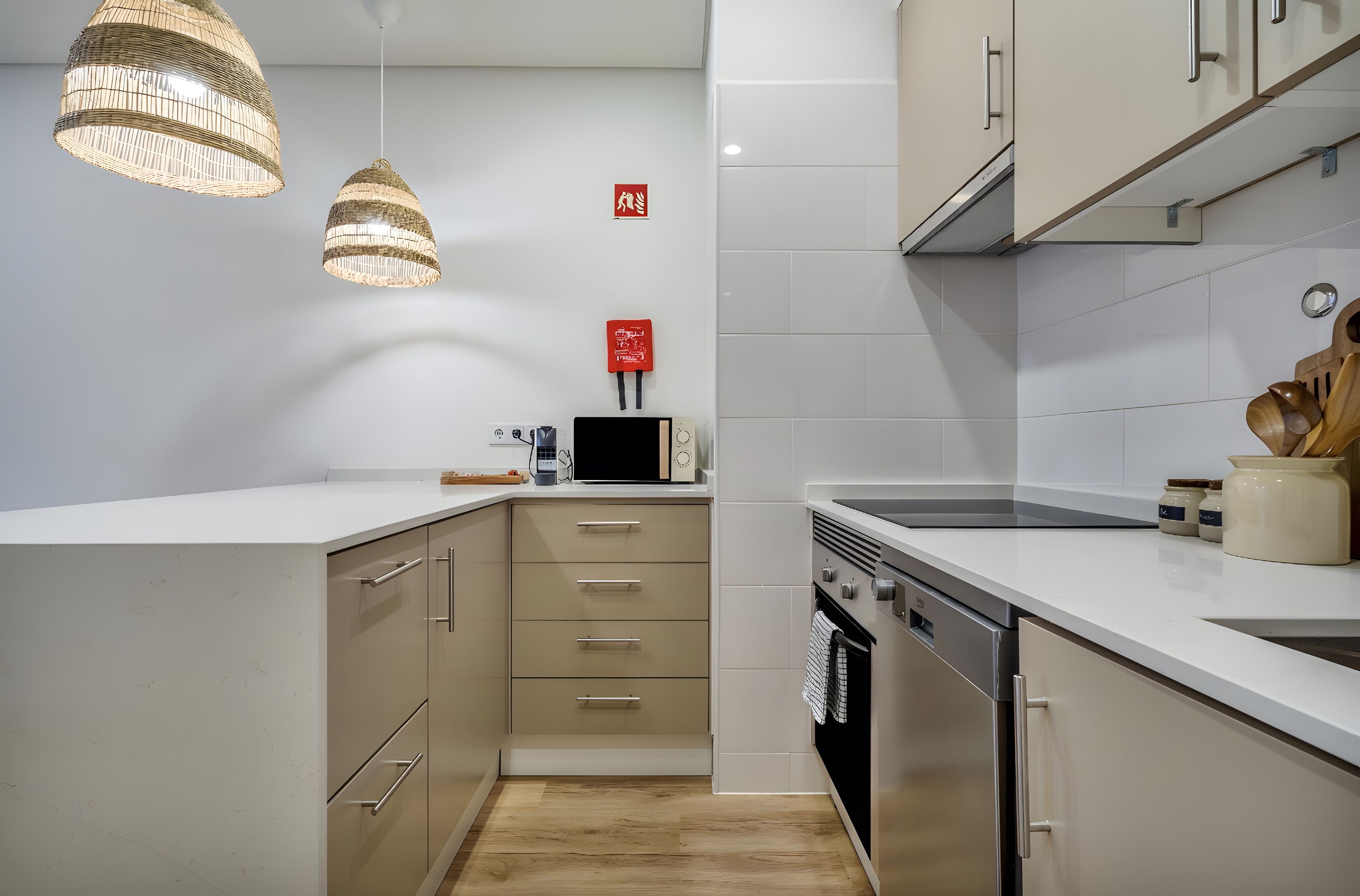A modern kitchen with white cabinetry, a built-in oven, and a microwave. There are two pendant lights and a red fire safety sign on the wall.