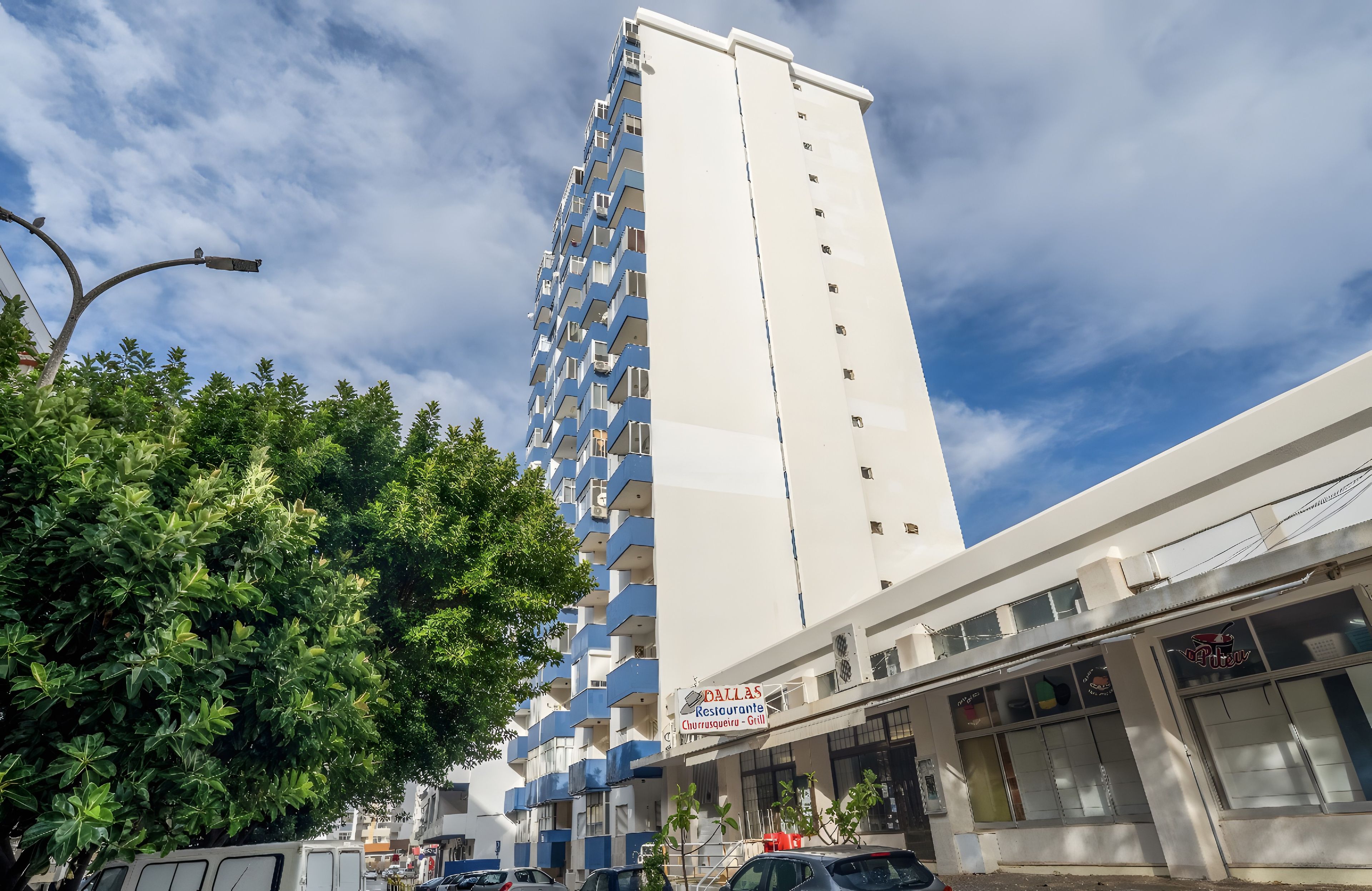 Un edificio residencial alto con balcones, una calle con coches estacionados y un restaurante llamado 'Dallas Restaurante Churrasqueiro - Grill' con mesas al aire libre.
