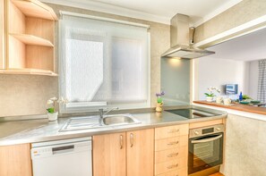 A modern kitchen with a dishwasher, oven, and a window with a leaf-patterned curtain. - Bella Italia Apartment near Playa de Alcudia (Alcúdia)