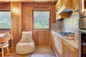 Ein Holzinnenraum mit einer Küche, einem Fenster mit Blick auf einen Wald, einem Stuhl und einem Tisch.
