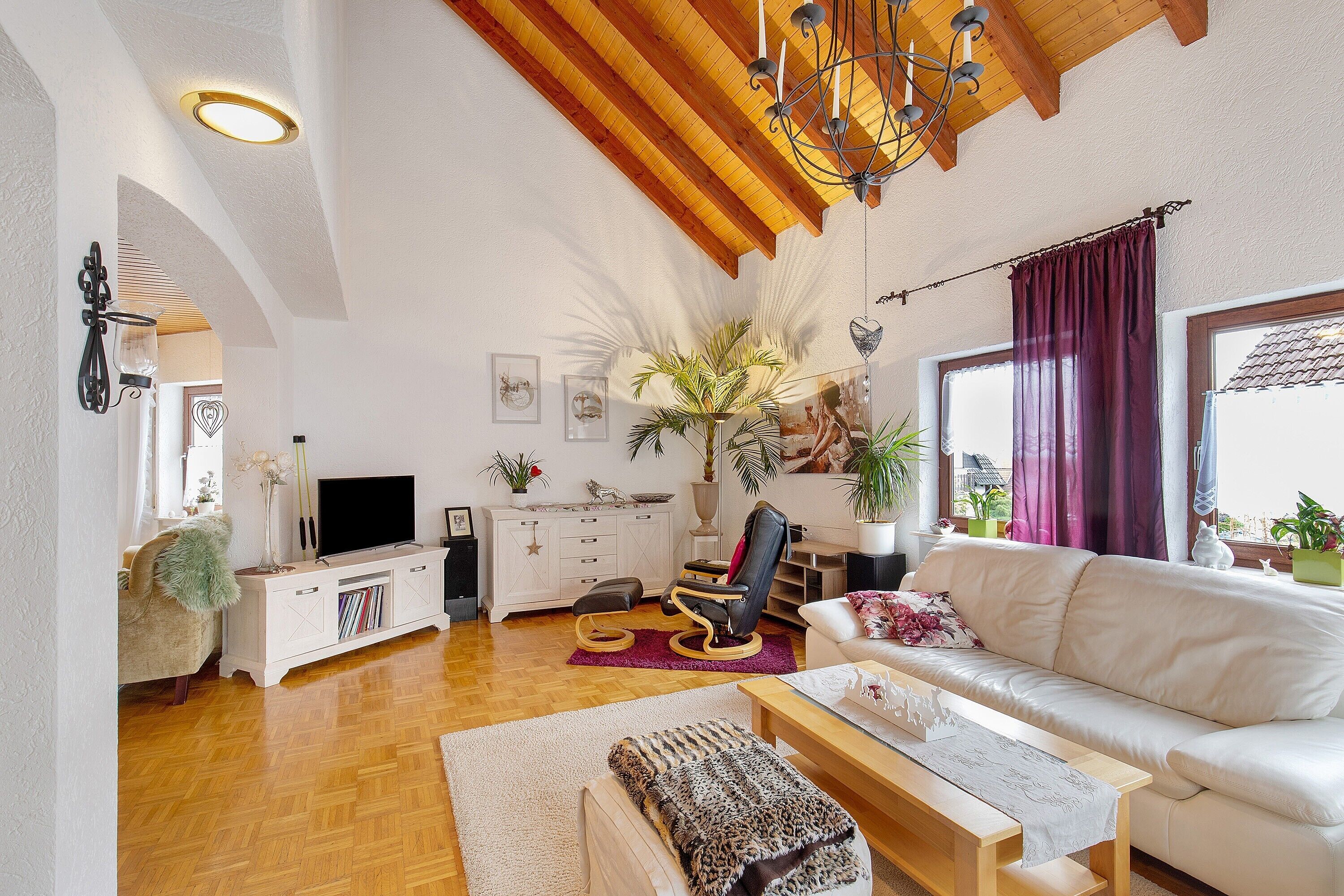 Ein Wohnzimmer mit einem weißen Sofa, Holzboden, einem Fernseher auf einem weißen Schrank, einem Deckenventilator und einem großen Fenster mit Vorhängen.