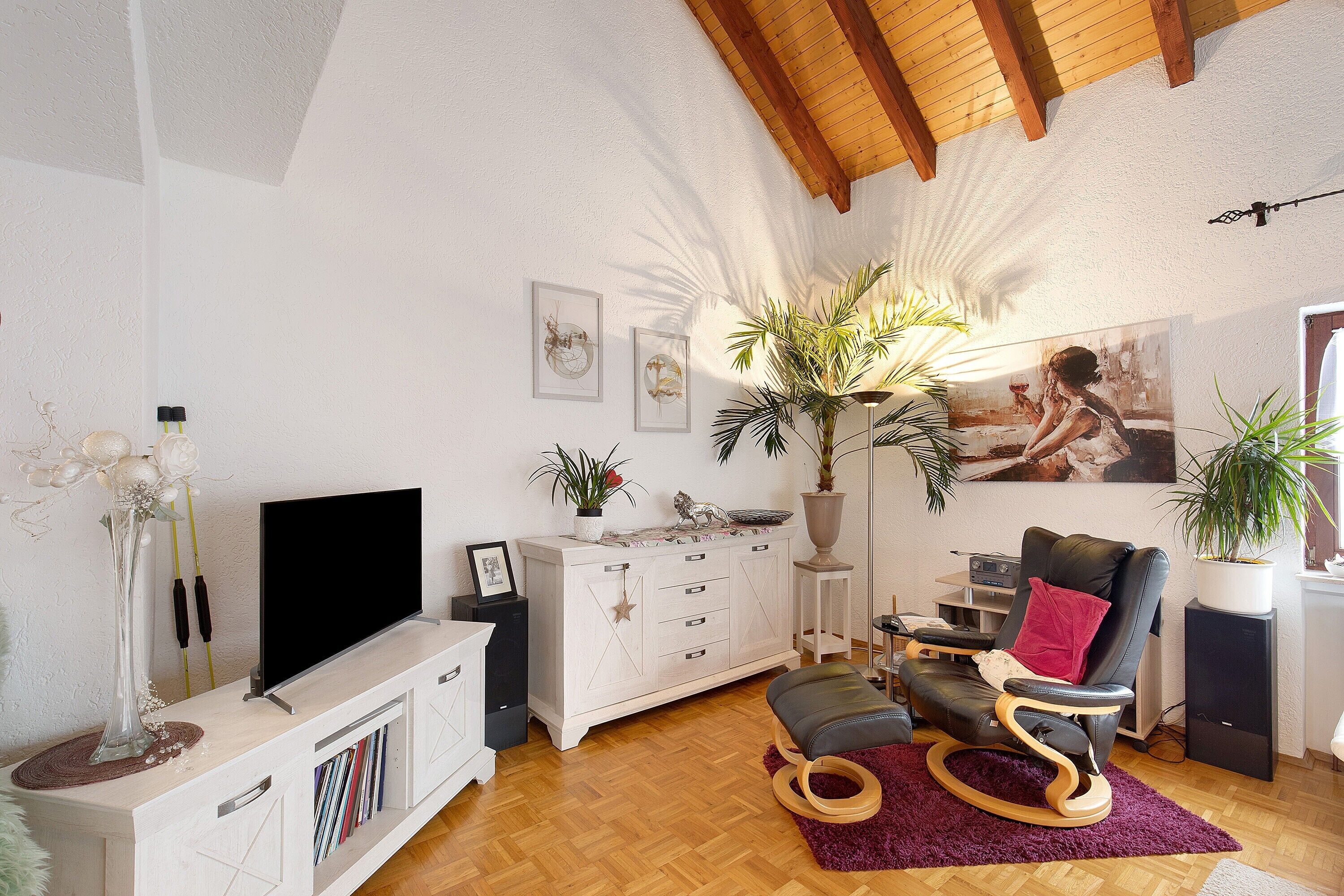 Een woonkamer met een televisie, een wit kastje, een leren fauteuil, potplanten en een groot schilderij.