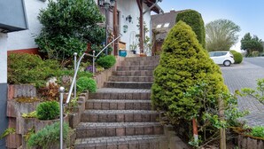 Uma casa residencial com uma escadaria, um carro estacionado na entrada e diversas plantas ao redor.